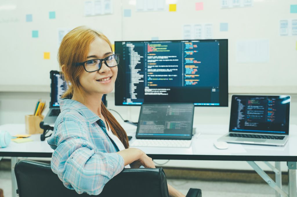 Young woman sitting at a desk with multiple monitors displaying code, smiling at the camera in a tech-savvy workspace, representing a remote medical coding role.