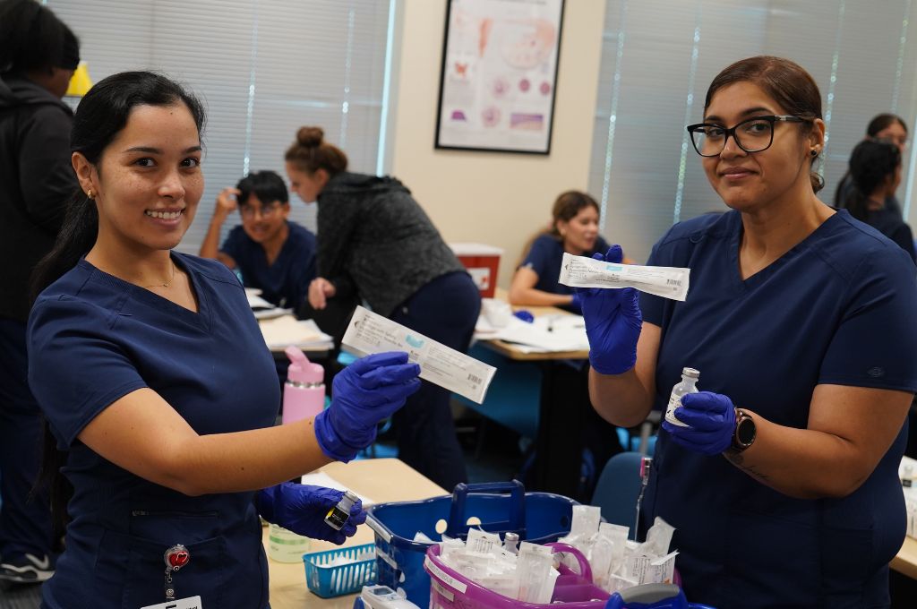 Two medical assistant students in California hold up syringes during hands-on training in a classroom setting.